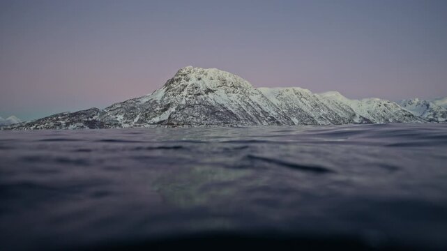 Split-level Arctic fjord with snowy Norway mountains beneath pastel twilight sky, golden hour alpenglow backdrop