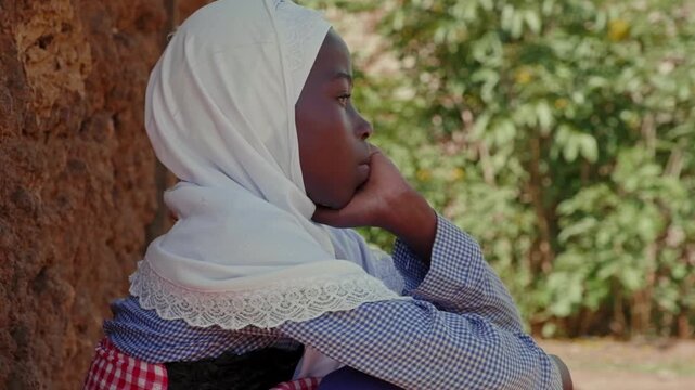 Close-up portrait of a thoughtful African girl in a white headscarf sitting beside a mud wall, showing quiet emotion, childhood and authentic rural village life.