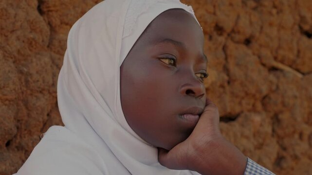 Close-up portrait of a thoughtful African girl in a white headscarf sitting beside a mud wall, showing quiet emotion, childhood and authentic rural village life.