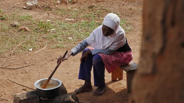 African child cooking in a metal pot over an open fire outside a mud house, showing family support, daily chores, food preparation and rural village life.