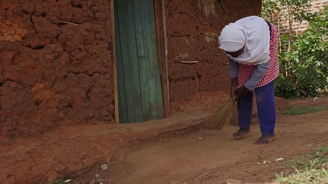African girl sweeping the ground outside a traditional mud house, showing daily household chores, family support and authentic rural village life in Africa.