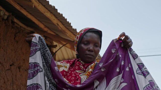 African woman hanging washed clothes outside a traditional mud house, showing laundry work, water scarcity, daily chores and authentic rural village life.