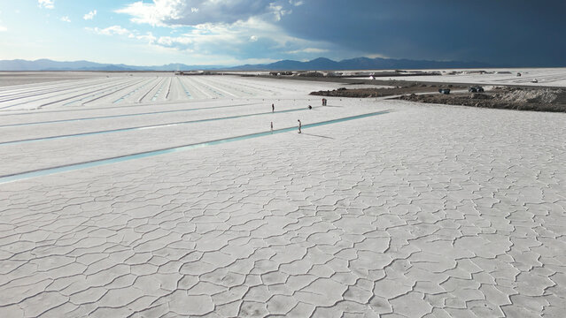 Aerial view of the Salinas Grandes salt flats with geometric crust patterns, turquoise water channels, and people walking under a dramatic sky in Tumbaya, Jujuy, Argentina.