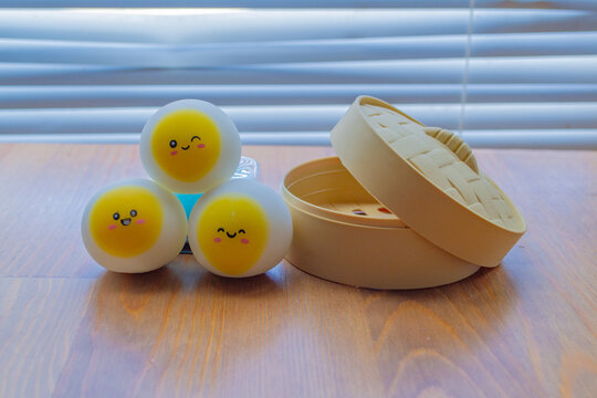 Three funny anti-stress dumpling squishies with cute smiley faces stacked in a neat pyramid on a wooden surface. Soft light through blinds in the background.