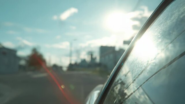 Car window reflecting a strong sun flare while driving on an urban street with blurred traffic lights and utility poles under a blue sky, conveying travel and journey