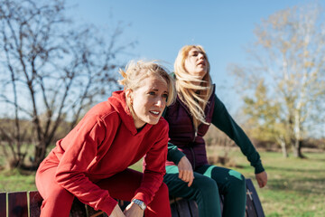 Obraz premium Two tired female friends in sportswear resting on a wooden bench, catching their breath after an intense outdoor running workout in a sunny park.