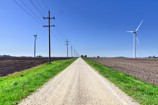 An unpaved road splices between telephone poles and a wind turrbine while stretching to and vanishing at the horizon. 
