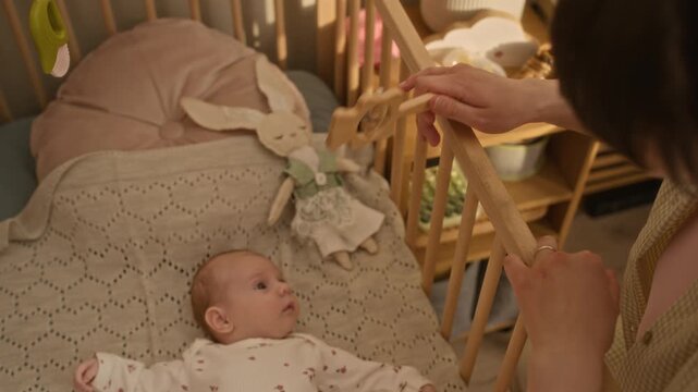 High angle handheld shot of unrecognizable mother comforting Caucasian infant baby girl lying in wooden crib with rattle toy in cozy nursery room