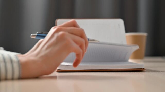 A person writes in a notebook using a pen while seated at a table. This moment highlights the process of note-taking and journaling, with a coffee cup nearby