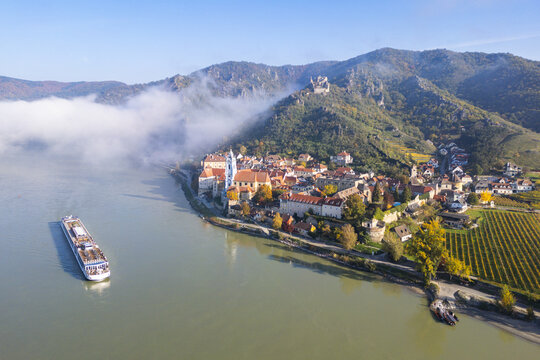 Aerial view of the historic town in the Wachau valley with its blue church tower, a cruise ship on the Danube river, and mist over the hills Durnstein, Lower Austria, Austria.