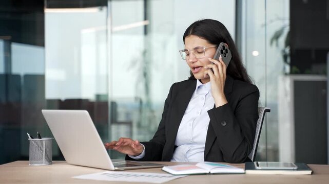 Busy businesswoman multitasking talking on smartphone and working on laptop while sitting in modern office workplace. Smiling female employee having business calls, manages tasks, using computer.