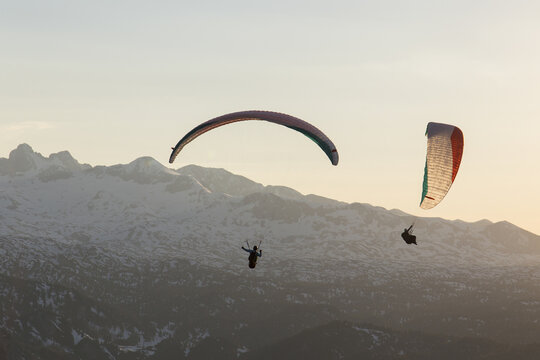 Aerial view of paragliders soaring over the snow-capped peaks of Hoher Dachstein during a soft sunset in Liezen District, Styria, Austria.