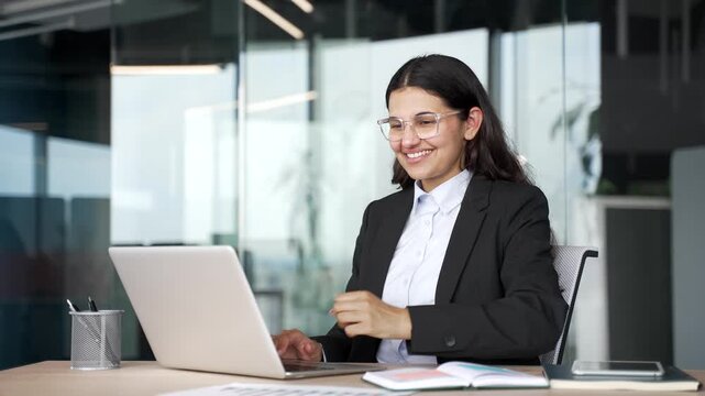 Smiling young businesswoman using laptop and reading good news in modern business office. Happy female employee reacting to positive online message, approval or successful business update