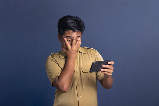 Indonesian civil servant man peeking through fingers at smartphone with shocked or scared face