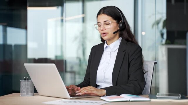Smiling woman corporate operator talking in headset working in customer support service on helpline telesale using laptop in business office. Young female agent of call center advises clients online.
