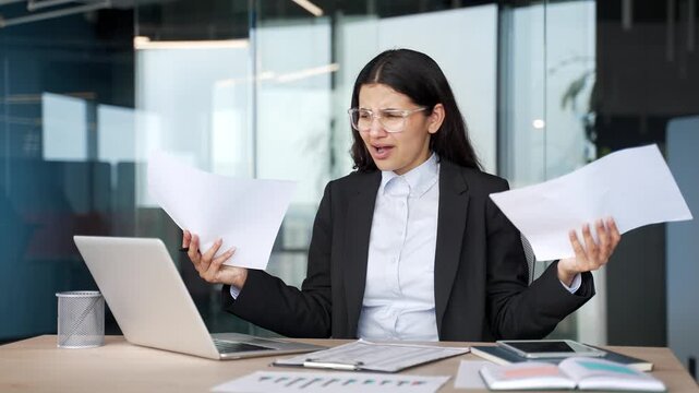 Confused young businesswoman holding documents and having difficulties with paperwork, reports and data. Puzzled female accountant struggle with business tasks while sitting at desk in office.
