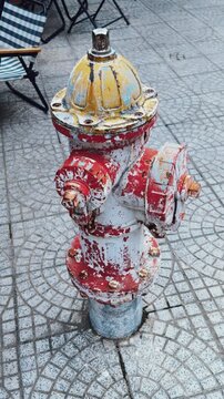 Worn fire hydrant standing on patterned pavement, showing peeling paint in red, white, yellow, and blue, representing urban infrastructure, aging, and street utility