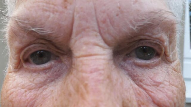 Close up wrinkled face of old grandmother looking into camera with a sad sight. Portrait of mature woman strongly screwing up her gray eyes. Sorrow facial expression of senior lady. Slow motion