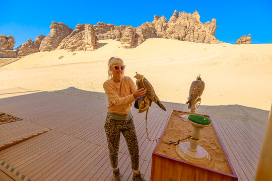 woman holding a peregrine falcon, resting on gloved hand, showcasing a desert tradition in Wadi Alna'am, AlUla, against a backdrop of rock formations and a luxury desert resort terrace in Saudi Arabia