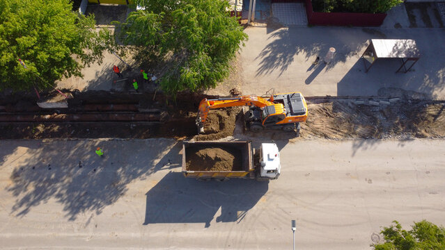 A high-angle drone shot capturing an orange hydraulic crawler excavator digging a trench and loading soil into the back of a white tipper dump truck