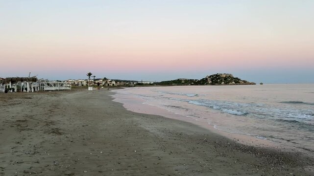 Sunset at sandy beach with gentle waves and distant buildings, suitable for a tranquil background