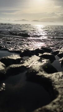 Morning sun reflecting on ocean water splashing over mossy volcanic rocks. Traditional boats float on the horizon beneath misty mountains, with soothing wave audio. Papuma Beach, Jember, Indonesia.