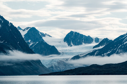 View of Lilliehookbreen glacier flowing between jagged mountains with low clouds and calm water in Svalbard, Svalbard and Jan Mayen.