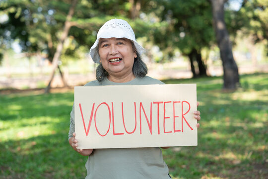 A senior woman smiles while holding a VOLUNTEER sign in a sunny park. The image highlights active aging, community service, and the spirit of giving back through outdoor social engagement