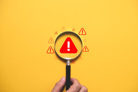 Warning alert concept with magnifying glass focusing on danger symbol over yellow background. Risk management, security monitoring, problem detection, caution and safety awareness concept.