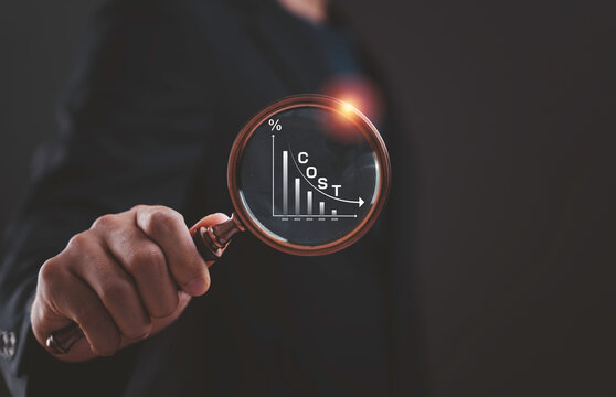 Businessman analyzing cost reduction trend through magnifying glass with declining financial graph. Expense control, budget optimization, business strategy and economic downturn concept.
