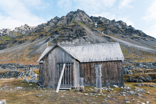 View of a rustic wooden cabin at Varsolbukta with a massive rocky mountain peak and sparse arctic tundra in Svalbard, Svalbard and Jan Mayen.