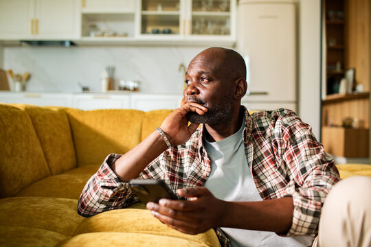 Thoughtful man holding smartphone on sofa in cozy living room