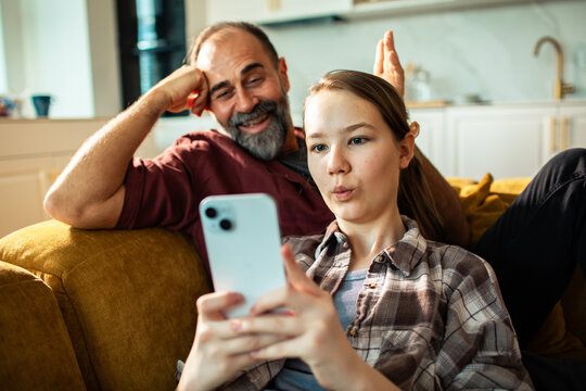 Father and teenage daughter laughing at smartphone on sofa at home