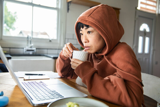Focused adult in hoodie drinking coffee while working from home kitchen