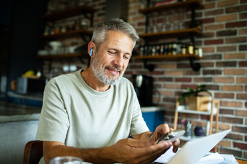 Fototapeta premium Smiling senior man using smartphone while working on laptop at home kitchen