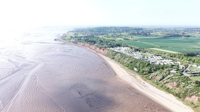 A steady 4K aerial view following the beach edge with smooth water patterns forming across the shoreline under warm seasonal brightness.