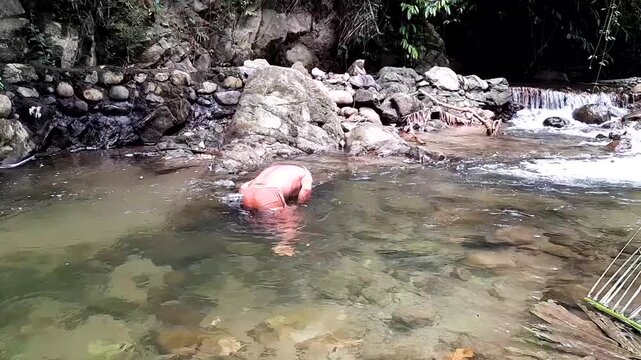 A man diving and swimming in a clear rocky river to catch fish using a traditional spear or bow mechanism.
