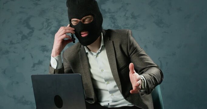 Scammer in balaclava talks on phone near laptop during online fraud call. Man gestures with hand as pressuring victim while managing deceptive scheme at desk