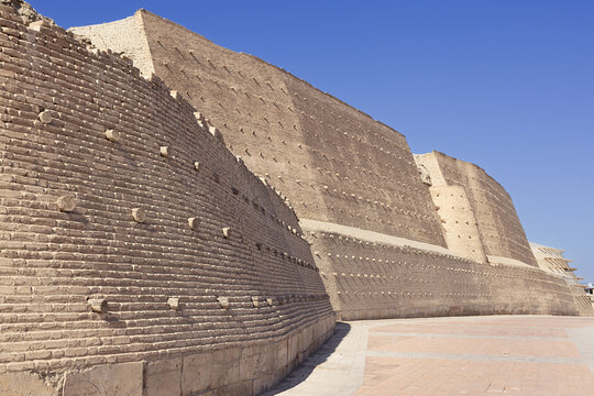 Massive fortress wall of the Ark of Bukhara with mud-brick construction and wooden beam supports under clear blue sky. Historic citadel architecture ideal for travel publications and heritage.