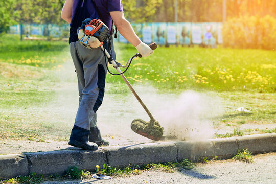 Municipal worker operating gas powered string trimmer to cut overgrown grass and weeds along concrete street curb during sunny day in summer. Professional landscaping and lawn maintenance service