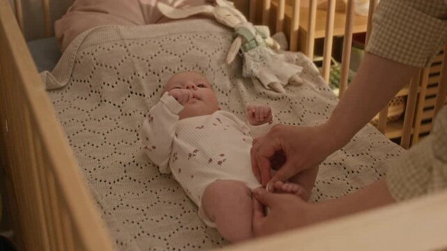 High angle slowmo shot of hands of unrecognizable mother dressing Caucasian baby girl lying in wooden crib in soft white onesie while changing clothes at home