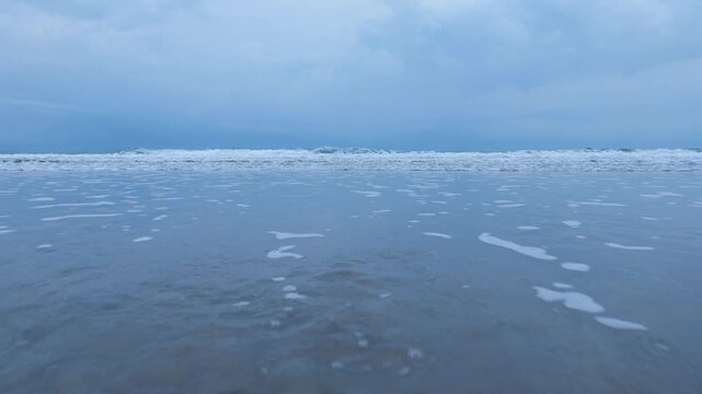 Cinematic footage of large, powerful ocean waves crashing forcefully onto a wide reflective sandy beach under a dark and stormy sky. Multiple wave lines roll in succession toward the shore