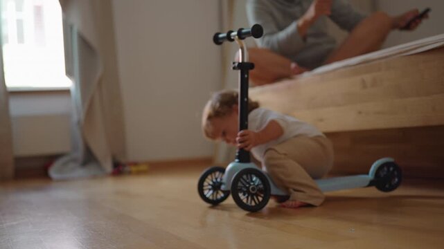 Toddler learning to ride small scooter indoors while repeatedly falling and getting back up capturing resilience, balance development and authentic childhood play at home