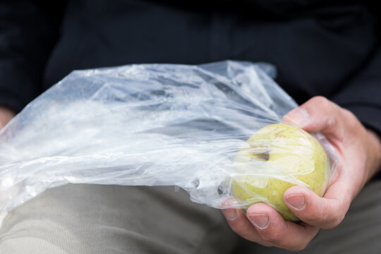 A man sitting outdoors holding a green apple partially covered by a crinkled plastic bag, conceptualizing consumer waste and environmental health.