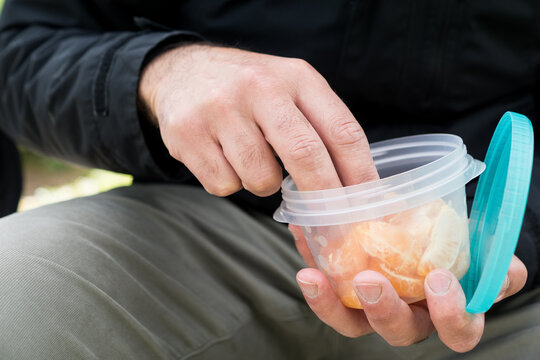 Close-up of a man's hand reaching inside a clear plastic tupperware container filled with orange mandarin segments to grab a healthy snack.