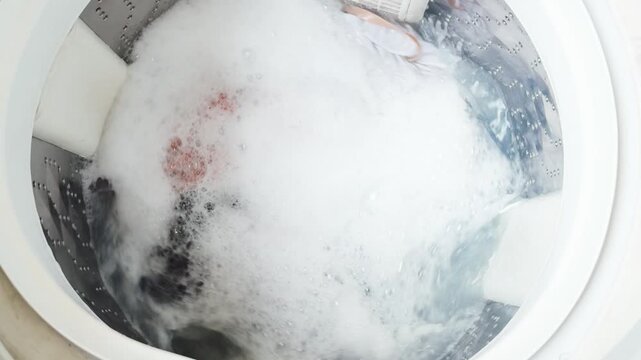 Washing machine completing a cycle, spinning dirty clothes with a powerful jet of water mixed with detergent, creating abundant white foam and bubbles inside the appliance drum