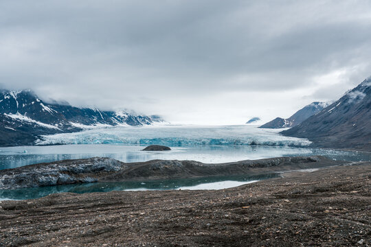 View of a massive glacier flowing into a fjord surrounded by snow-capped mountains and rocky terrain under an overcast sky Svalbard and Jan Mayen.