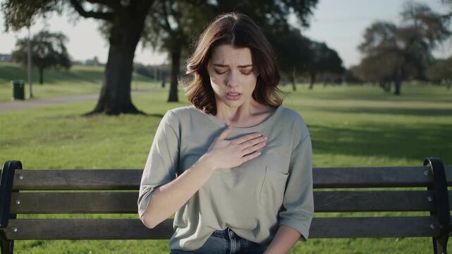 Young caucasian woman clutching chest outdoors, college student on park bench gasping for breath, hand pressed to heart, anxious expression, sunlight through oak trees, green grass background, subtle