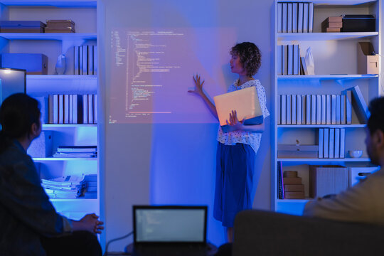 A professional East Asian female software engineer stands holding a laptop and gesturing toward lines of code projected on a wall display, presenting data scripts to her multiethnic colleagues during 