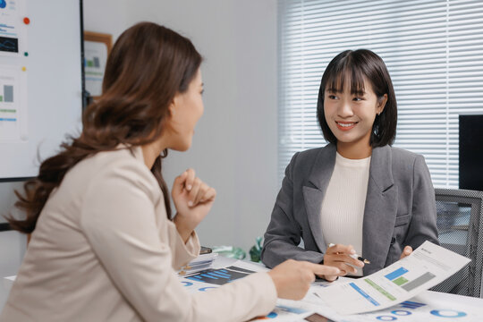 Two professional Asian businesswomen collaborating, analyzing charts and graphs, and planning new projects in a modern office setup